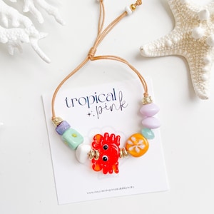 May include: A colorful beaded bracelet with a crab charm and the text "tropical + pink". The bracelet features a red crab, orange fruit, and various pastel-colored beads. The adjustable cord is tan.