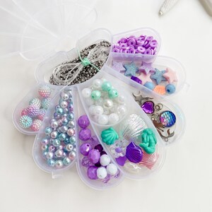 May include: A clear plastic clamshell container filled with various beads, charms, and string for making jewelry. The container is divided into sections, each containing a different type of bead or charm. The beads are in shades of purple, blue, green, and white. The charms include seashells, stars, and a mermaid tail.