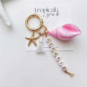 May include: A pink seashell charm with a gold starfish, a white pearl, and a white tassel attached to a gold key ring. The charm also has white beads spelling out "MERMAID" with gold lettering.