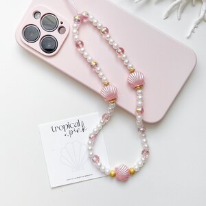 May include: A pink and white beaded phone charm with a pink seashell charm. The charm is attached to a pink phone case with a white background. The text "tropical + pink" is visible on a white card.