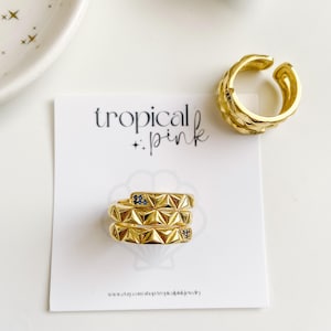 May include: Gold-toned jewelry set featuring a textured ear cuff and two stacked rings with geometric designs and small blue accents. The rings and ear cuff are displayed on a white surface with a card that reads "tropical pink".