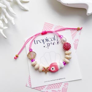 May include: A pink and white beaded bracelet with a tropical theme. The bracelet features a pink cord, a seashell charm, a starfish charm, and a pink evil eye charm. The card behind the bracelet says "tropical pink".