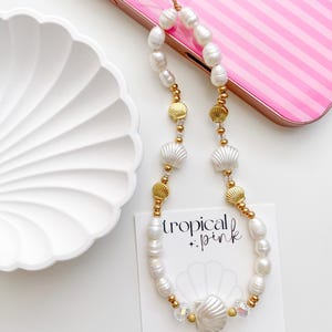 May include: A phone charm featuring white freshwater pearls, gold-toned metal shell beads, white shell-shaped beads, and clear faceted crystals. The charm is attached to a display card with the text tropical + pink and a shell graphic.