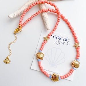 May include: A coral-colored beaded necklace with gold seashell and starfish charms. The necklace features a gold clasp and a small crab charm. The text "tropical pink" is visible on a white card.