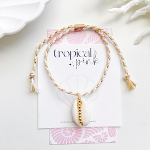May include: White and gold braided necklace with a cowrie shell pendant. The necklace features a white and gold cord with a tassel at each end. The pendant is a white cowrie shell with gold beads. The text "tropical pink" is on the card.