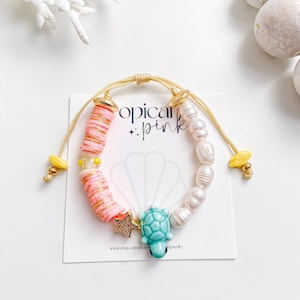 May include: A handmade bracelet featuring pink and gold beads, freshwater pearls, and a turquoise turtle charm. The bracelet includes a gold star charm and is strung on a gold cord. The text "tropical + pink" is visible on a card.