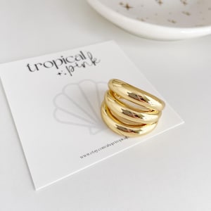 May include: Gold ring with four bands stacked on top of each other. The ring is on a white surface with a white card that says "tropical pink" and a website address.