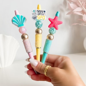 May include: Three colorful pens with beach-themed toppers. One pen has a seashell, another has a starfish, and the third has the text "I need vitamin sea". The pens are pink, yellow, and teal, with gold and glitter accents.