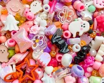 May include: A collection of assorted craft beads in various shapes and colors, including white ghosts, pink smiley faces, clear flowers, black bats, and orange stars. The mix features translucent, opaque, and iridescent finishes in a vibrant palette.