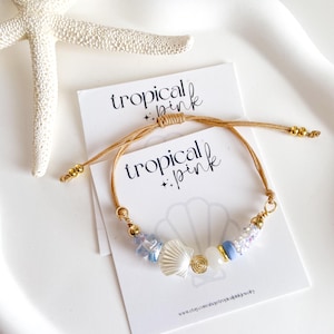 May include: A tan cord bracelet featuring a collection of beads including a white shell, a gold spiral, a light blue bead, and a clear iridescent bead. The bracelet is displayed on a white card with the text tropical + pink printed in black.