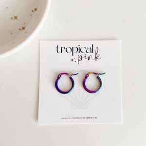 May include: A pair of rainbow iridescent hoop earrings on a white card with the text "tropical : pink" and the website address "www.etsy.com/shop/tropicalpinkjewelry".