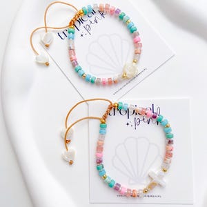 May include: Two colorful beaded bracelets with the text "tropical pink" on the cards they rest on. The bracelets feature a variety of pastel-colored beads, gold accents, and decorative charms. One bracelet has a white rose charm, and the other has a white cross charm.