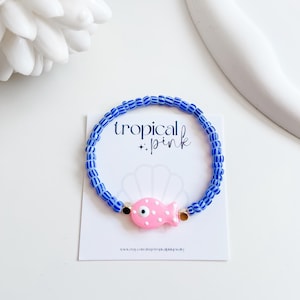 May include: A blue beaded bracelet with a pink fish charm. The bracelet features a series of blue and white striped beads. The pink fish charm has a white eye and gold accents. The words "tropical + pink" are printed on the card.