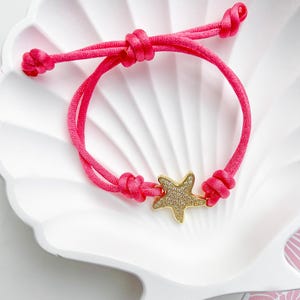 May include: A bright pink adjustable bracelet with a gold-colored starfish charm. The starfish is encrusted with small, clear crystals. The bracelet is displayed on a white, seashell-shaped dish. The cord is knotted at each end.