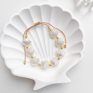 May include: A delicate bracelet featuring white seashell charms and gold-colored beads strung on a tan cord. The bracelet is displayed on a white, shell-shaped dish, creating a coastal or beach-themed aesthetic.
