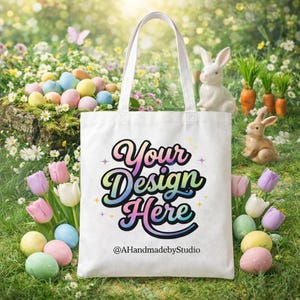 May include: A white tote bag with the text "Your Design Here" in a colorful, retro font. The bag is set against a spring scene with Easter eggs, bunnies, and flowers. The text "@AHandmadebyStudio" is below the main text.