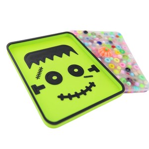 Può includere: Due custodie rettangolari per telefono. Una è verde lime con un design a volto di Frankenstein nero. L'altra è trasparente con perline colorate e una piccola zucca. Le custodie sono probabilmente per uno smartphone e a tema Halloween.