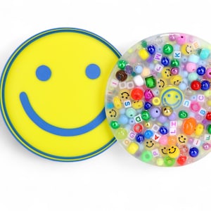 Può includere: Due sottobicchieri rotondi. Uno è giallo con una faccina sorridente blu. L'altro è trasparente con perline colorate, faccine sorridenti e perline con lettere. I sottobicchieri sono su sfondo bianco.