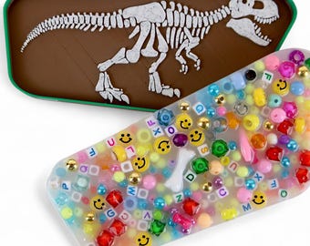 May include: Two rectangular cases. One is brown with a white dinosaur skeleton. The other is clear with colorful beads, smiley faces, and letter beads. A fun, playful design.