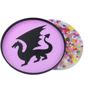 Può includere: Un sottobicchiere rotondo di colore viola con una silhouette nera di un drago. Il sottobicchiere è reversibile e l'altro lato è trasparente con perline colorate incorporate nella resina.