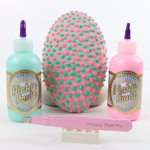 以下が含まれることがあります： 「Hand Crafted Picky Paint Made in USA」と金色に書かれたラベルが付いた、ターコイズブルーとピンクの塗料のボトルが2本。中央には、ピンクとターコイズブルーのドットがついたグレーの卵型のオブジェクトがあります。手前に、ピンクのピンセットと「Picky Party」と書かれた白いレゴブロックがあります。