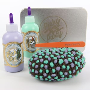 Könnte beinhalten: Zwei Flaschen Farbe mit violetten Deckeln und goldenen Etiketten, auf denen "Picky Paint" und "Hand Crafted Made in USA" steht. Eine silberne Dose mit dem Text "Picky Party" darauf. Ein braunes, violettes und grünes, gesprenkeltes, eiförmiges Objekt.