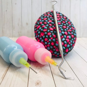 Op de afbeelding: Twee kleine flesjes verf met gele puntjes en een metalen stippenhulpmiddel. De flesjes zijn blauw en roze. Een zwarte steen met roze en blauwe stippen is op de achtergrond.