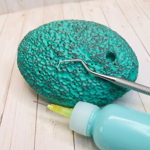 Könnte beinhalten: Ein türkisfarbenes Keramik-Ei mit einer strukturierten Oberfläche und mehreren Löchern. Neben dem Ei liegen ein kleines Metallwerkzeug und eine Flasche türkisfarbener Farbe.