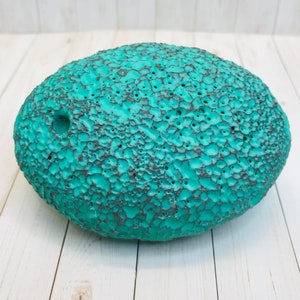 Op de afbeelding: Een ovaalvormig, getextureerd keramiekobject in tealblauw met een gat aan de zijkant. Het object heeft een ruw, onregelmatig oppervlak.