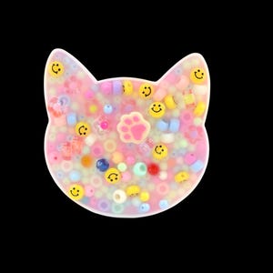 Può includere: Un pezzo di resina a forma di gatto riempito di perline colorate e dettagli di faccine sorridenti. Il design include un'impronta di zampa rosa al centro. Le perline sono di vari colori, tra cui rosa, giallo, blu e bianco. Lo sfondo è nero.