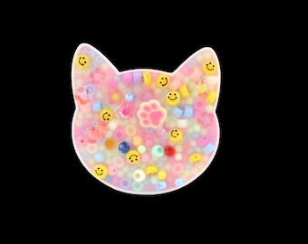May include: A cat-shaped resin piece filled with colorful beads and smiley face accents. The design includes a pink paw print in the center. The beads are in various colors, including pink, yellow, blue, and white. The background is black.