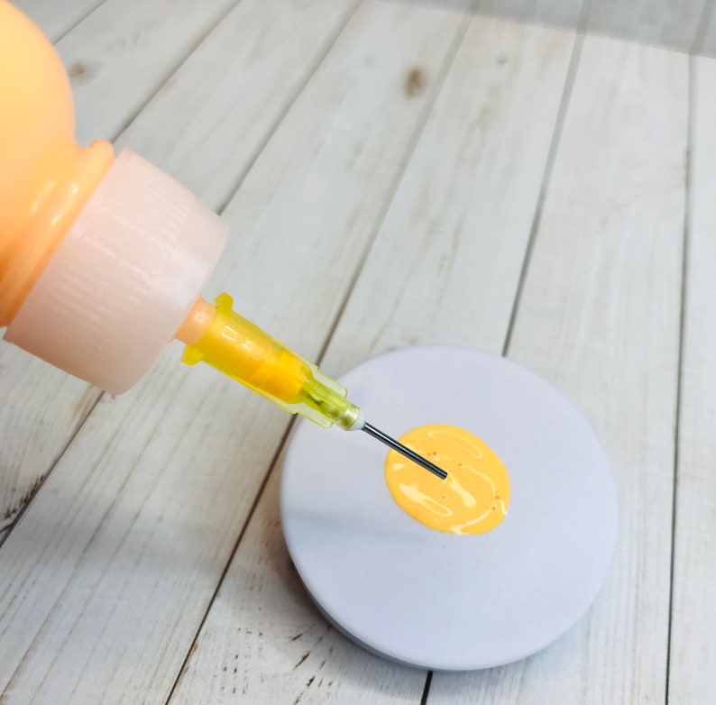 May include: A yellow paint bottle with a needle attached to the tip, dispensing a small amount of yellow paint onto a round, light gray surface.