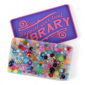 Può includere: Una scatola rettangolare in resina trasparente piena di perline colorate, lettere e piccole perline color oro. Un coperchio viola con finiture rosa e il testo "Support your local LIBRARY" è sopra la scatola.