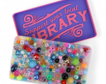 May include: A rectangular, clear resin box filled with colorful beads, letters, and small gold-colored beads. A purple lid with pink trim and the text "Support your local LIBRARY" is above the box. The beads are various shapes and sizes.