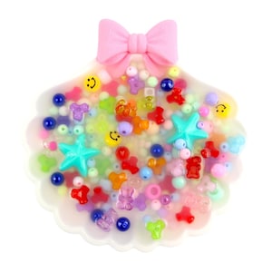 Pode incluir: Uma peça artesanal em forma de concha cheia de contas coloridas, estrelas e carinhas sorridentes. Uma fita rosa está presa no topo. As contas são de várias cores, incluindo azul, vermelho, amarelo e verde.