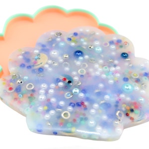 Puede incluir: Un joyero en forma de concha con una parte superior de resina translúcida azul claro llena de cuentas de colores, perlas y detalles plateados. El plato se asienta sobre una base de color melocotón con un borde festoneado verde menta.