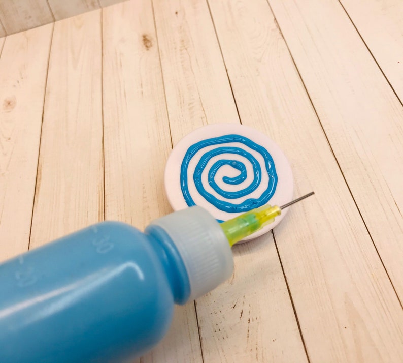 Op de afbeelding: Een wit keramiek bord met een blauw spiraalontwerp. Een blauwe fles met een gele punt wordt gebruikt om het ontwerp te maken.