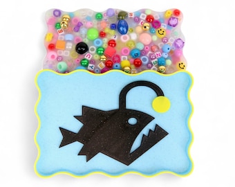 May include: A whimsical, layered art piece featuring a black anglerfish silhouette on a light blue background with a yellow border. The top layer is filled with colorful beads and small objects, creating a playful, textured effect.