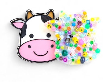 May include: Two cow-shaped pins. One is a cartoon cow face with a black outline, white face, pink nose, and black eyes. The other is a cow shape filled with colorful beads, smiley faces, and letters. The pins are on a white background.