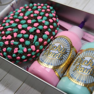 Op de afbeelding: Een set van twee flessen verf in een metalen doos. De flessen zijn gelabeld "Picky Paint" en zijn roze en turquoise. De doos bevat ook een bruin sponsje met roze en turquoise stippen.