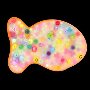 Pode incluir: Um projeto de artesanato em resina em forma de peixe preenchido com contas coloridas. As contas são de vários tamanhos e formas, incluindo quadrados, círculos e rostos sorridentes. A resina é de cor laranja translúcida e o fundo é preto.