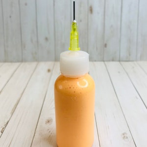 Op de afbeelding: Een klein, doorzichtig flesje met een witte dop en een gele naald die aan de bovenkant is bevestigd. Het flesje is gevuld met een heldere oranje vloeistof.