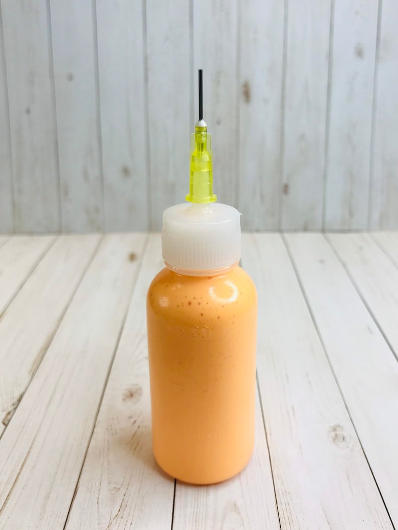 May include: A small, clear bottle with a white cap and a yellow needle attached to the top. The bottle is filled with a bright orange liquid.