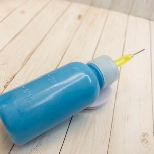 Op de afbeelding: Een blauwe plastic fles met een gele naald die aan de bovenkant is bevestigd. De fles heeft markeringen voor 10, 20 en 30 milliliter.