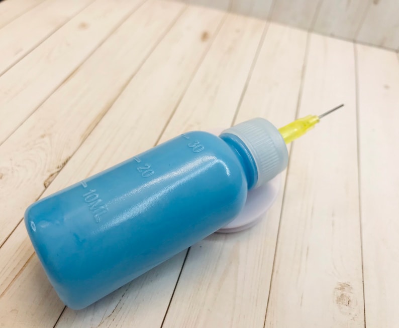 Op de afbeelding: Een blauwe plastic fles met een gele naald die aan de bovenkant is bevestigd. De fles heeft markeringen voor 10, 20 en 30 milliliter.