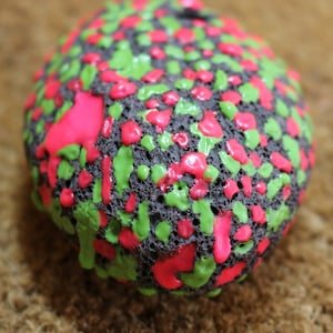 Op de afbeelding: Een ronde, donkergrijze steen bedekt met roze en groene verfspatten.
