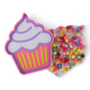 Può includere: Un contenitore a forma di cupcake rosa e viola con un motivo di glassa bianca e strisce verdi. Accanto, un contenitore trasparente a forma di nuvola pieno di perline colorate, faccine sorridenti e cubi con lettere. Su sfondo bianco.