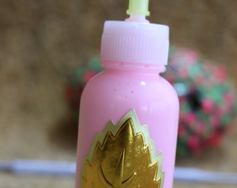 Puede incluir: Una botella rosa con una pegatina de hoja dorada y una punta de aplicador de plástico transparente. La botella está sobre una superficie marrón.