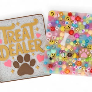 Può includere: Due sottobicchieri quadrati a tema animali domestici. Uno presenta il testo "TREAT DEALER" in oro con un'impronta di zampa marrone e cuori rosa. L'altro è pieno di perline colorate, faccine sorridenti e un osso.