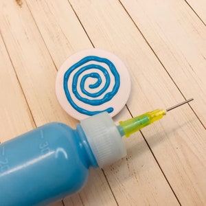 Op de afbeelding: Een witte keramische schijf met een blauw spiraalontwerp erop geschilderd. Een blauwe plastic fles met een gele punt wordt gebruikt om de verf aan te brengen.